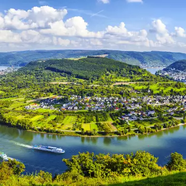 A wide shot of a bend in the Moselle River from it's surrounding hills under broad daylight, two river cruise ships can be seen creating gentle waves in the water.